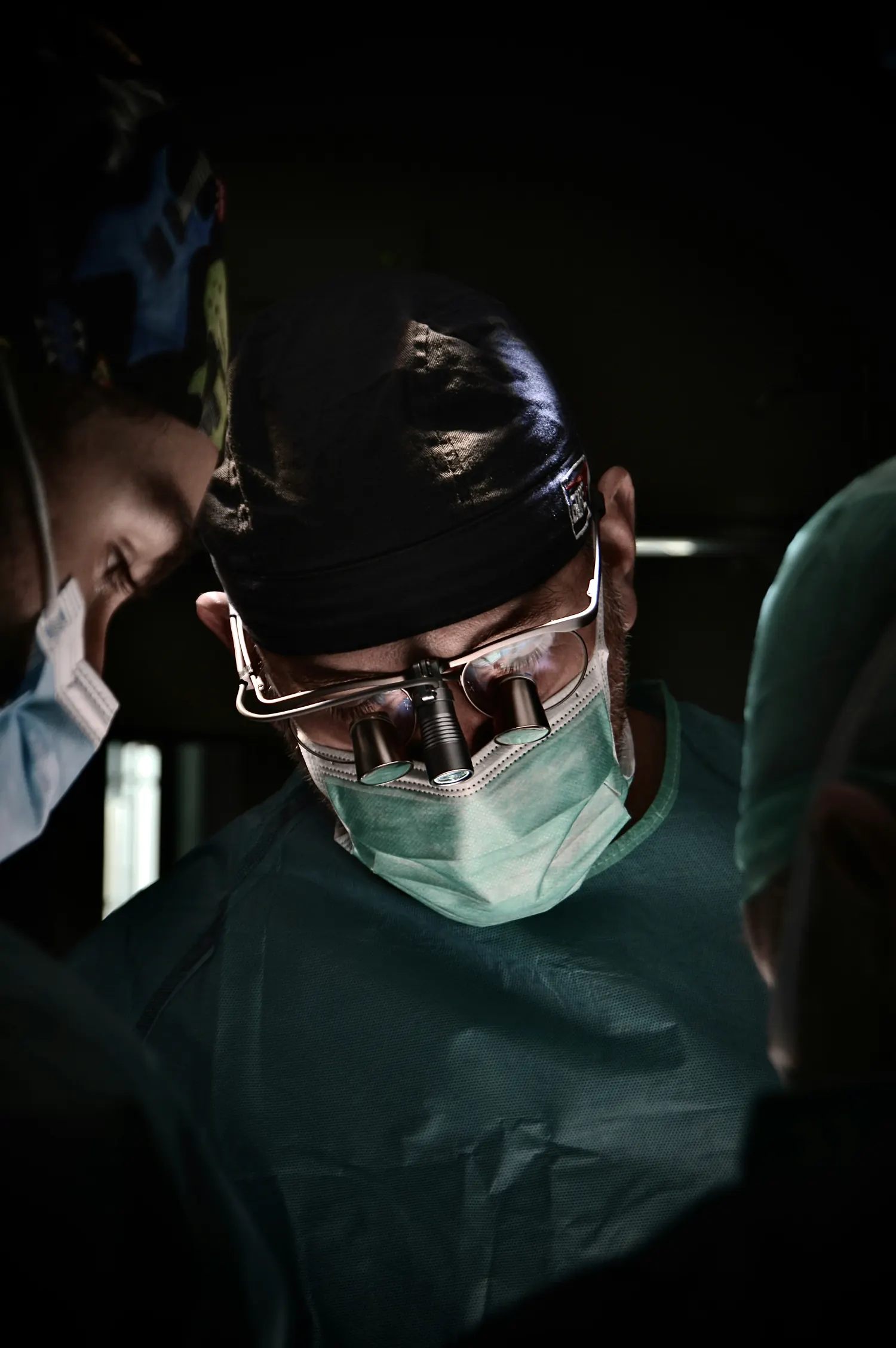Black and white photo of a surgeon in a mask, cap, and magnifying loupes, looking down with intense focus