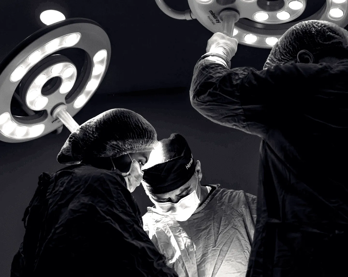 Black and white photo of surgeons in masks and gowns focused on a procedure under bright overhead operating lights