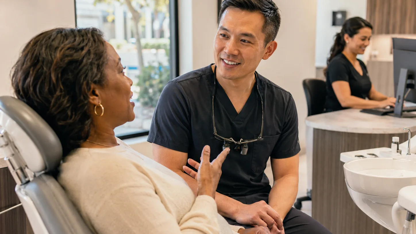 A dentist talks with a patient in the dental chair while a colleague works at the front desk
